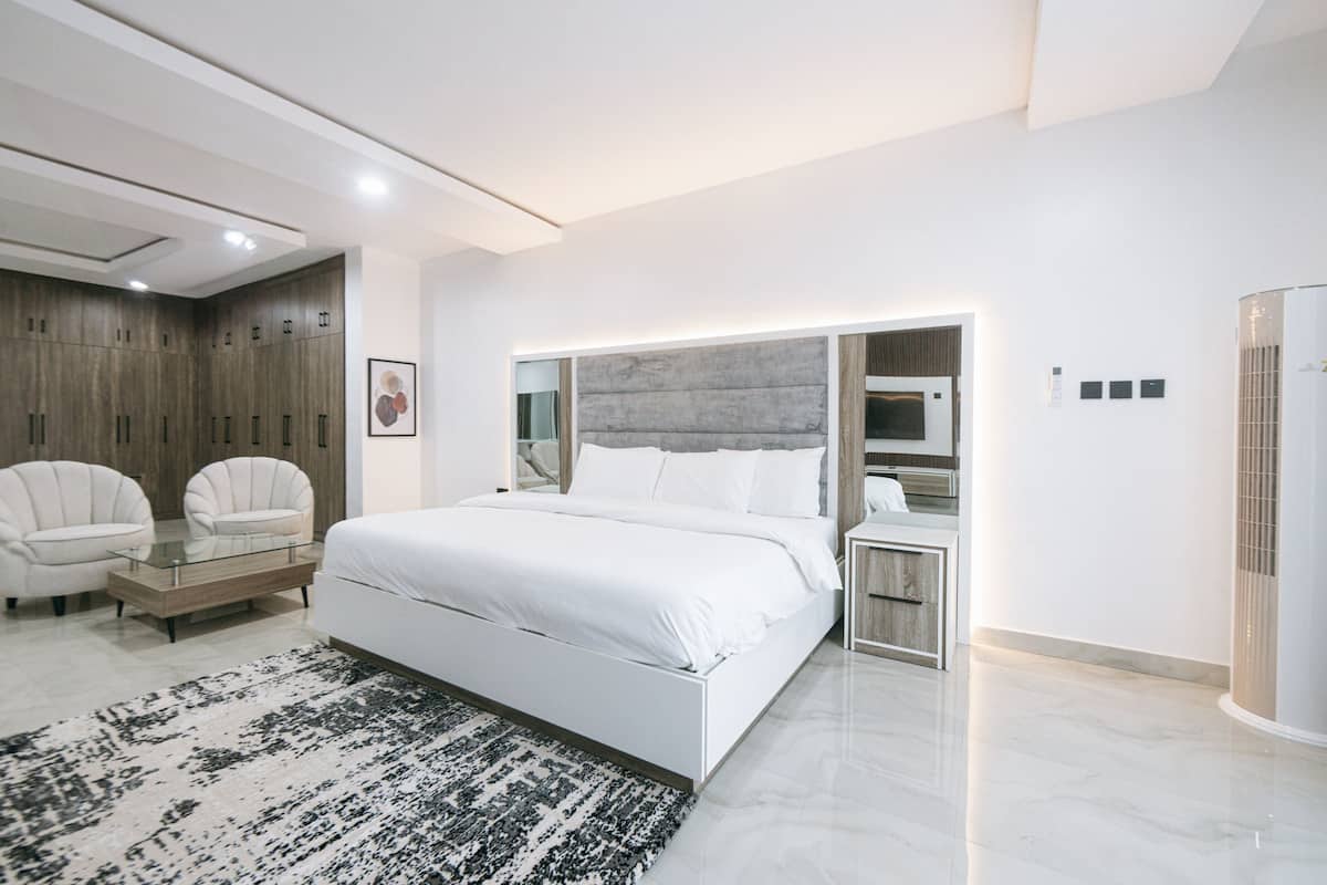 A modern bedroom with a large bed, a glass coffee table, two armchairs, a built-in wardrobe, and a bedside table with a lamp.