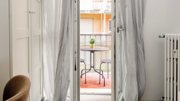 Eine geöffnete Tür, die auf einen Balkon mit Tisch, Stühlen und einem Blumentopf führt, mit Blick auf ein Gebäude mit orangenen Wänden.