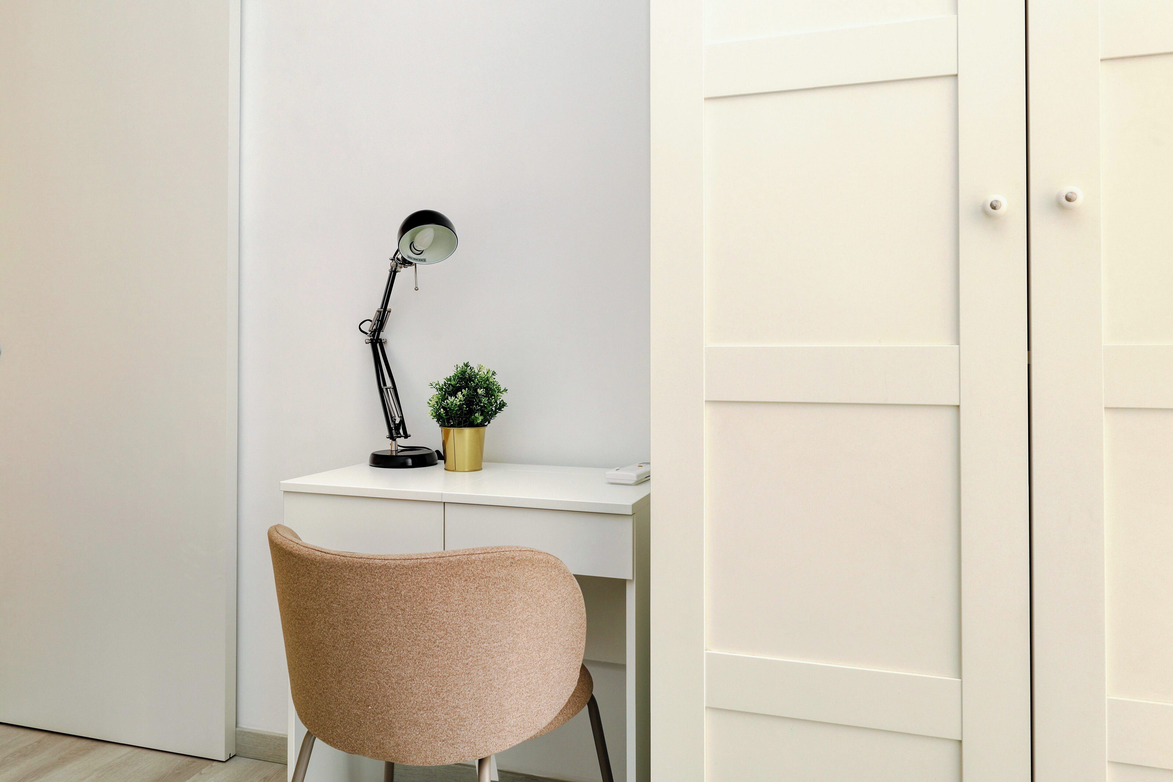 A white desk with a black desk lamp, a small potted plant, and a brown chair in a room with white walls and a white door.