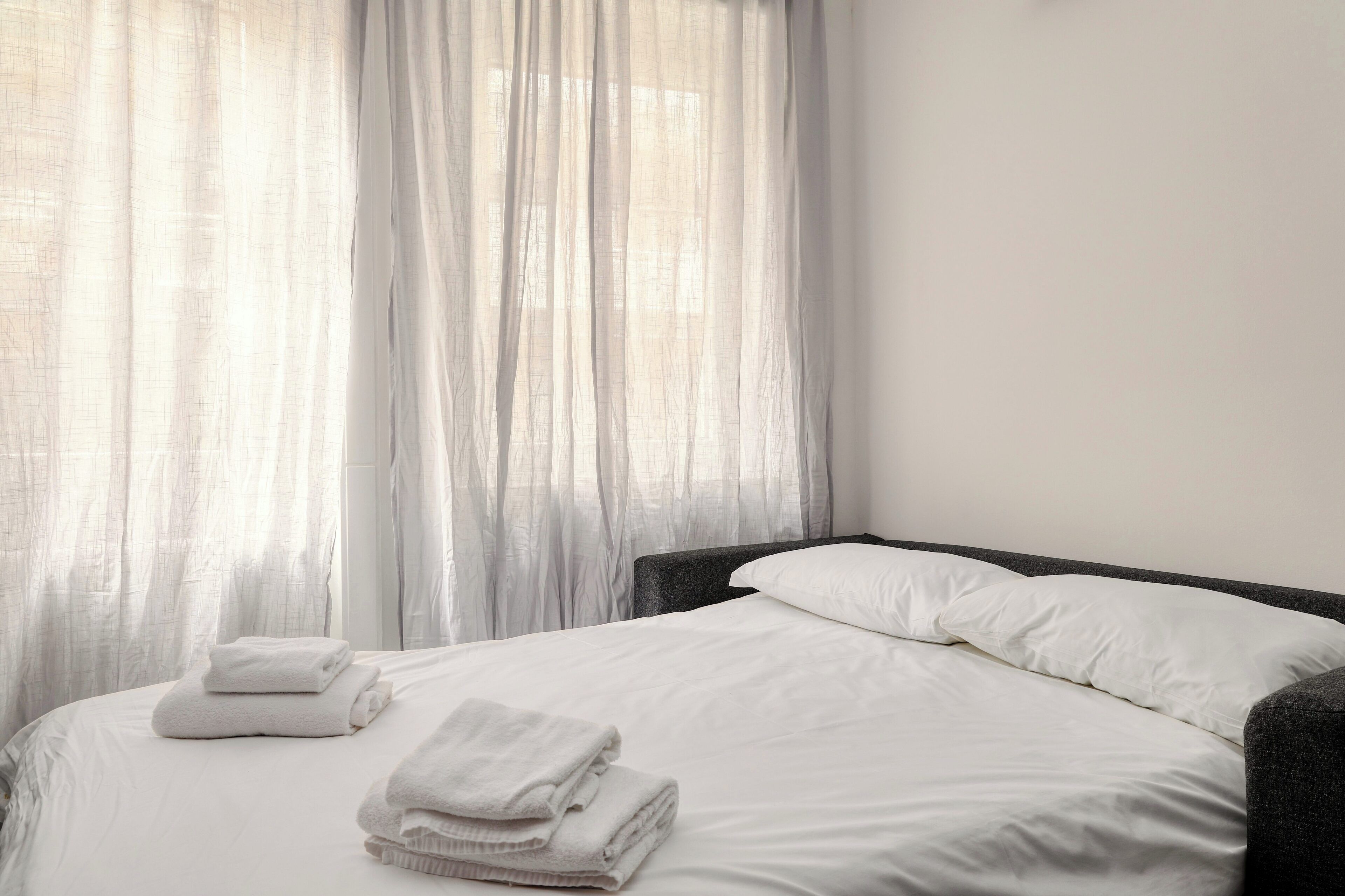 A neatly made bed with white linens and pillows, a dark headboard, and sheer white curtains.