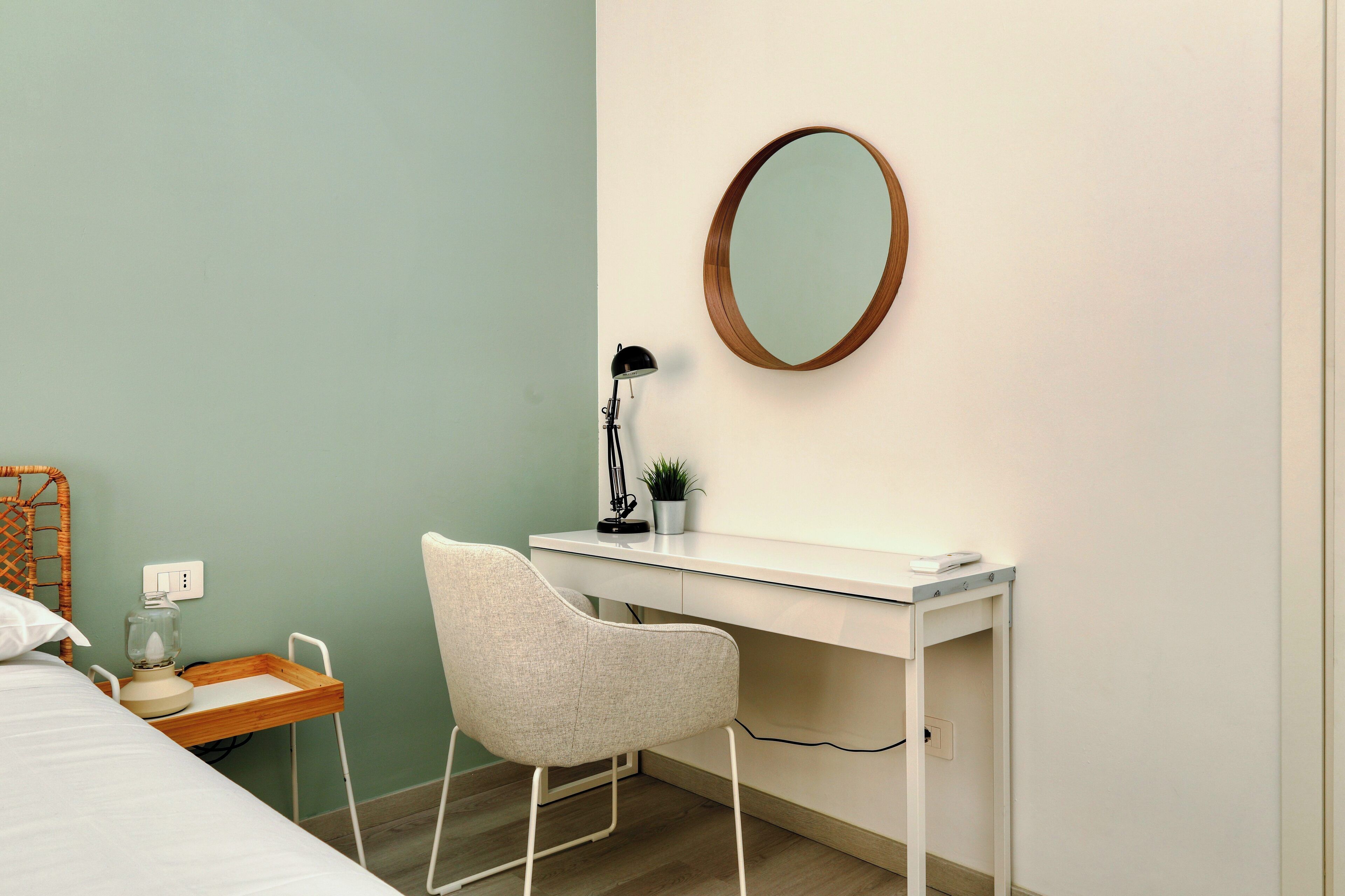 A bedroom with a bed, a desk, a chair, a mirror, a lamp, and a small plant.