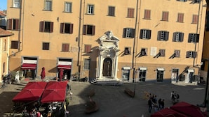 A courtyard with outdoor seating, a fountain, and a building with multiple windows and a statue. - Window To The Tower (Pisa)