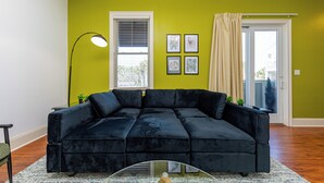 Un salon avec un canapé d’angle noir, une table basse en verre, un lampadaire et une fenêtre avec des stores.