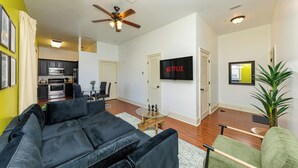 Un salon avec un canapé noir, un fauteuil vert, une table basse en verre et un ventilateur de plafond.