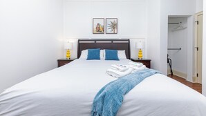Un lit bien fait, avec du linge de lit blanc et un plaid bleu, deux oreillers bleus et une tête de lit au fini foncé. On y trouve deux tables de chevet avec des lampes et un tableau encadré au-dessus du lit.