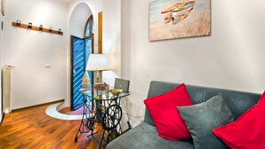 A modern living room with a grey sofa, red cushions, a glass coffee table, a laptop, and a lamp. There is a painting on the wall and a coat rack with hooks. - The Lake View (Castel Gandolfo)