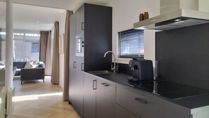A modern kitchen with dark cabinetry, a stainless steel sink, and a coffee machine. There is a window with blinds and a view of the outside.