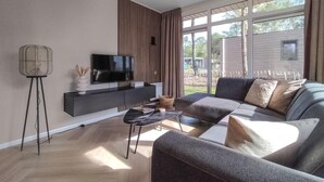 A modern living room with a large flat-screen TV on a black console, a grey sofa with multiple cushions, a small coffee table, and a tall floor lamp.