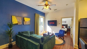 A modern living room with a green sectional sofa, a ceiling fan, a flat-screen TV, and a kitchen area visible through an open door.