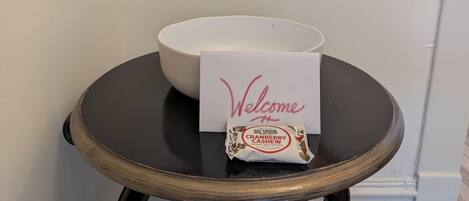A white bowl on a dark round table with a 'Welcome' card and a packet of 'Big Spoon Cranberry Cashew Peanut Butter Bar' beside it.