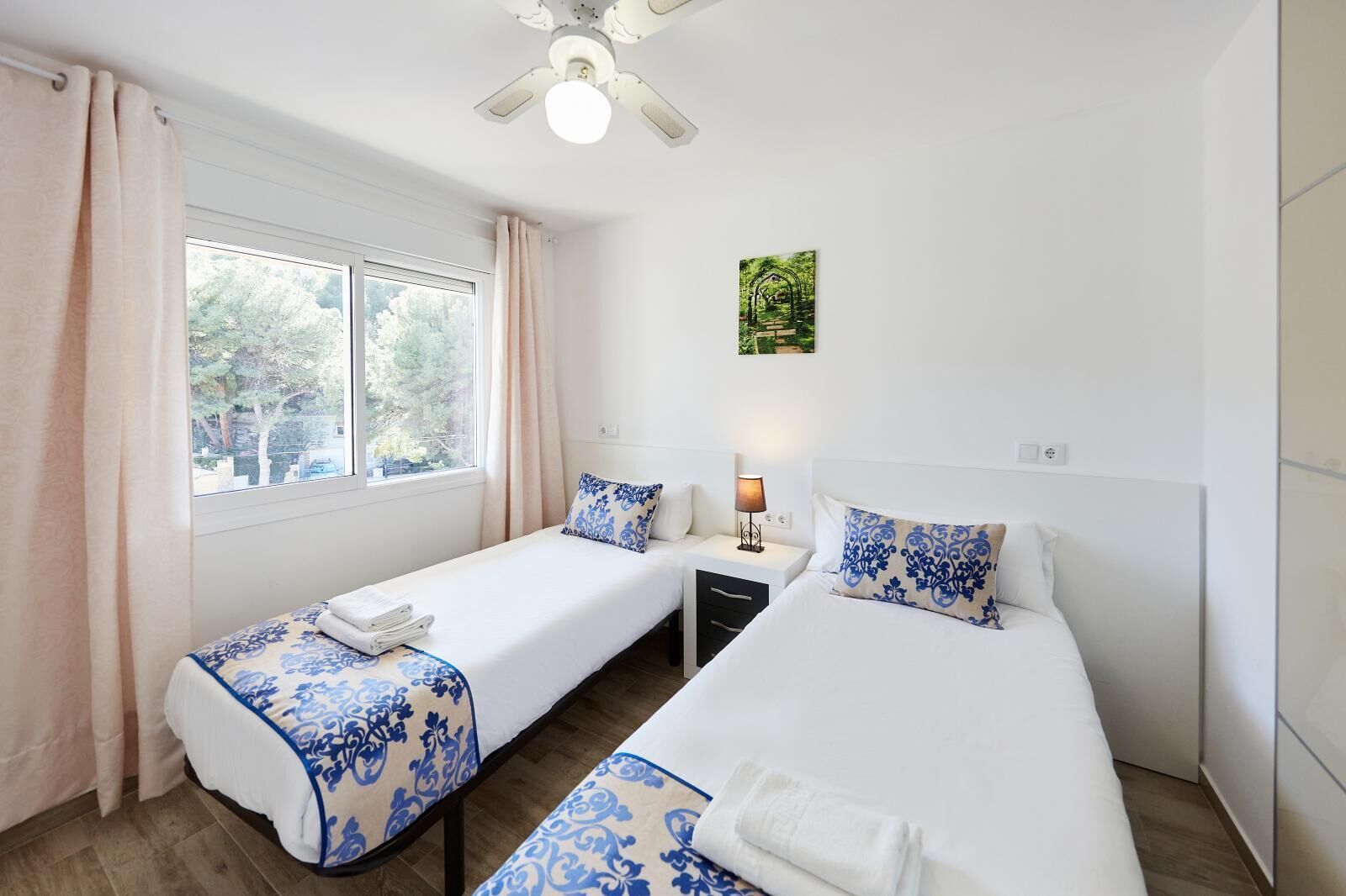 A hotel room with two beds, a window with curtains, a ceiling fan, and a wall-mounted lamp.