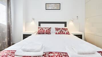 Una cama bien hecha con sábanas blancas y almohadas rojas estampadas, dos mesitas de noche con lámparas, y una obra de arte enmarcada en la pared.