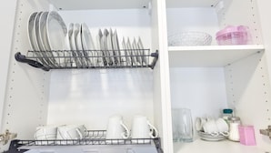Een keuken met een afwasschuur vol borden en kommen, een plank met bekers en een roze container, en een kast met potten en een glas.