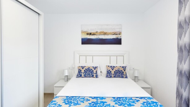 Een slaapkamer met een wit bed, twee blauw gemêleerde kussens, een wit hoofdeinde en een schilderij van een landschap boven het bed.