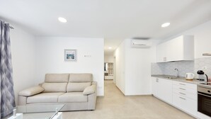 Een modern appartement met een beige bank, een glazen salontafel, een keuken met witte kasten en een betegelde spatwand, en een gang die naar een andere kamer leidt.