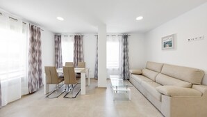 Een moderne woonkamer met een beige bank, een glazen salontafel en een eethoek met een witte tafel en stoelen.