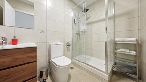 Een moderne badkamer met een douchecabine, een toilet, een wastafel met een rode zeepdispenser en een handdoekhouder met opgevouwen handdoeken.