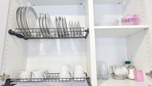 Een keuken met een afwasschuur vol borden en kommen, een plank met bekers en een roze container, en een kast met potten en een glas.