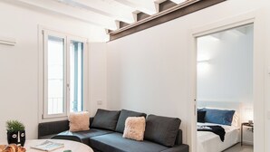 A modern, well-lit room with a grey sofa, a small round table with a cup and a plate, a bed with blue and white bedding, and a window with blinds. - Suite JOIVY Lake 11 (Como)