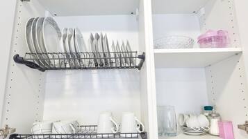 A kitchen with a dish rack holding plates and bowls, a shelf with cups and a pink container, and a cabinet with jars and a glass.