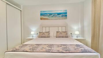 Un lit bien fait, recouvert d’une couvre-lit à motifs floraux, avec deux lampes de chevet et un tableau représentant une scène de plage au-dessus du lit.