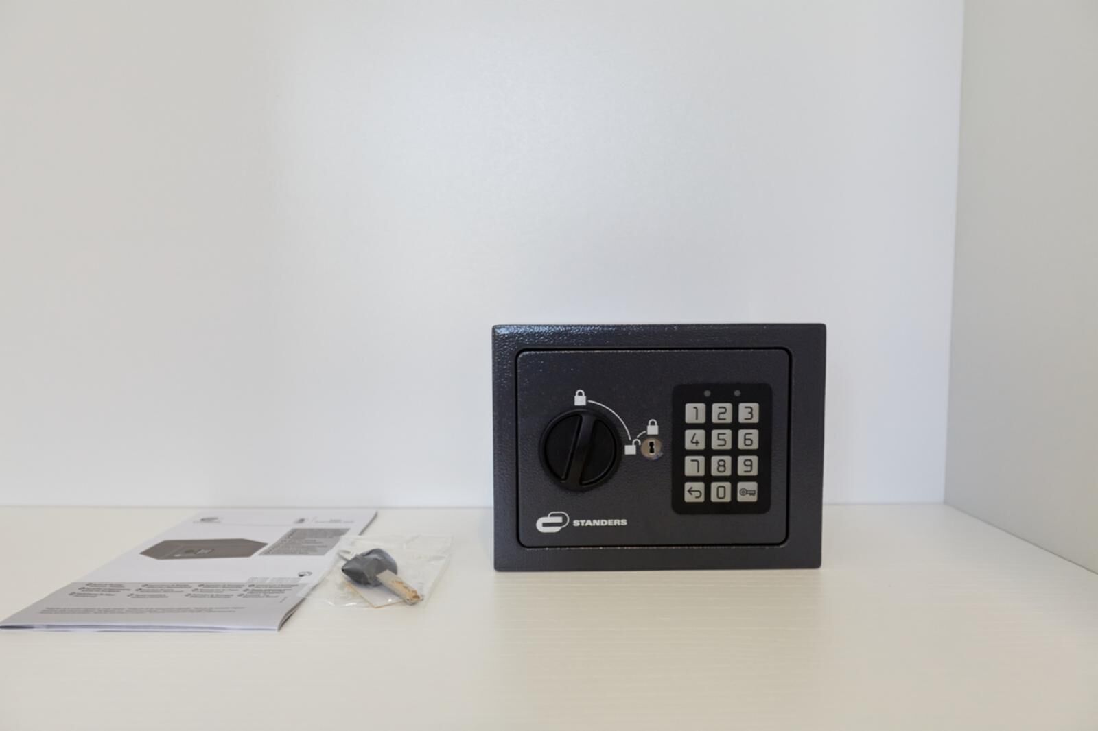 A black safe with a keypad and a keyhole, placed on a white surface with documents and a keychain nearby.