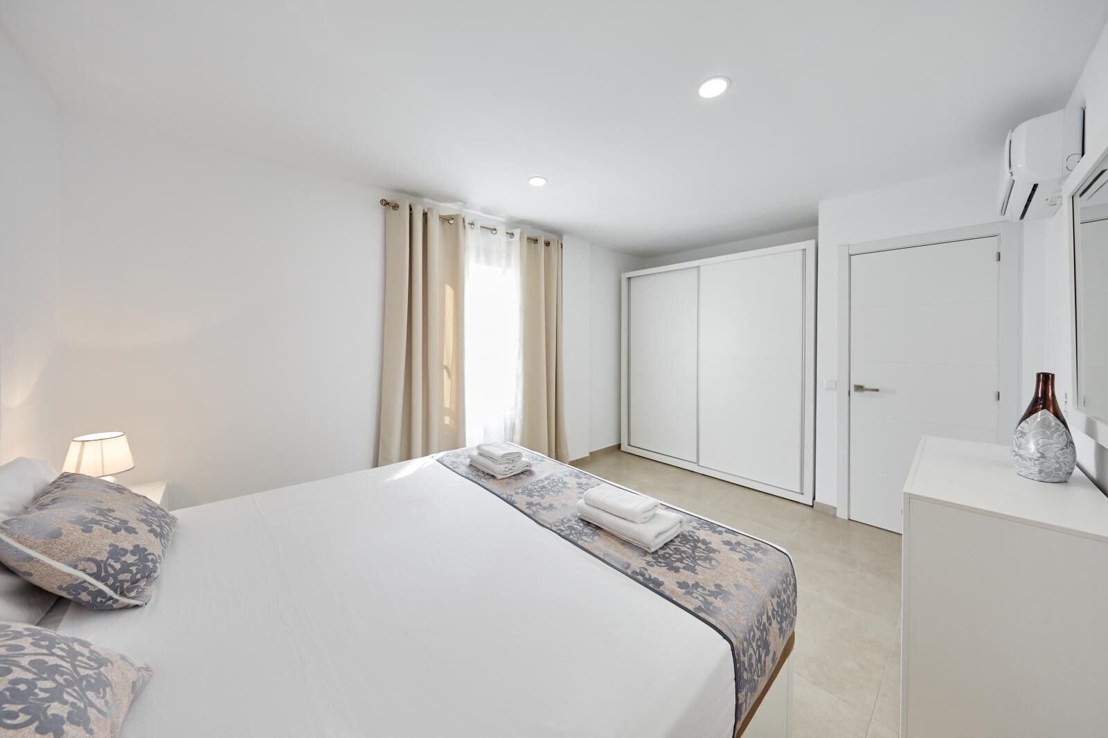 Una habitación de hotel moderna con una cama grande, una lámpara de noche, un armario y un florero sobre una cómoda.