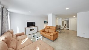 Un salon moderne avec un canapé, une table basse en verre et un téléviseur posé sur un meuble. On aperçoit à l’arrière-plan un coin repas avec des chaises et une table.