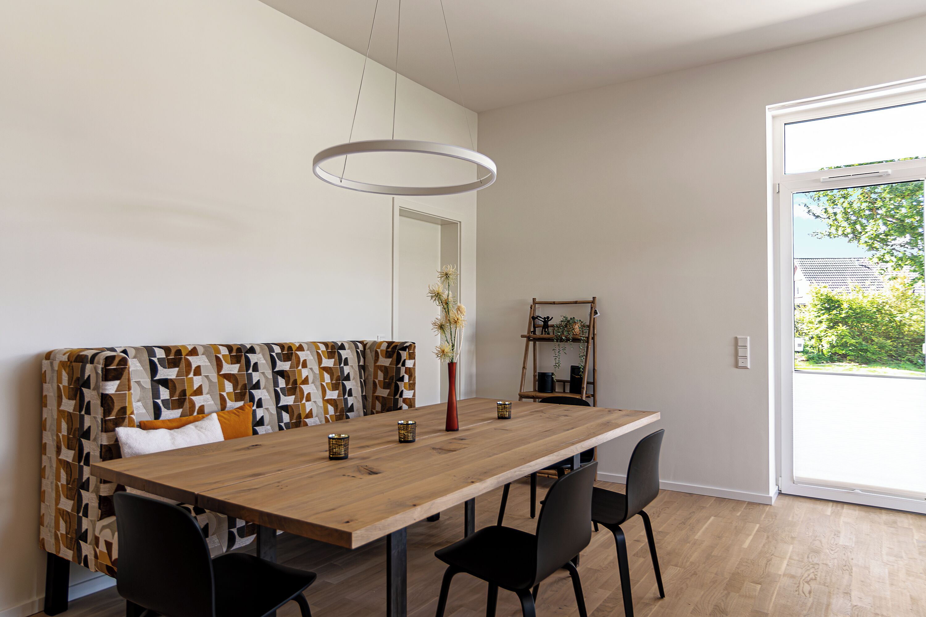Un coin repas avec une table en bois, des chaises noires, un banc à motifs, un vase de fleurs et une vue à travers une porte vitrée.