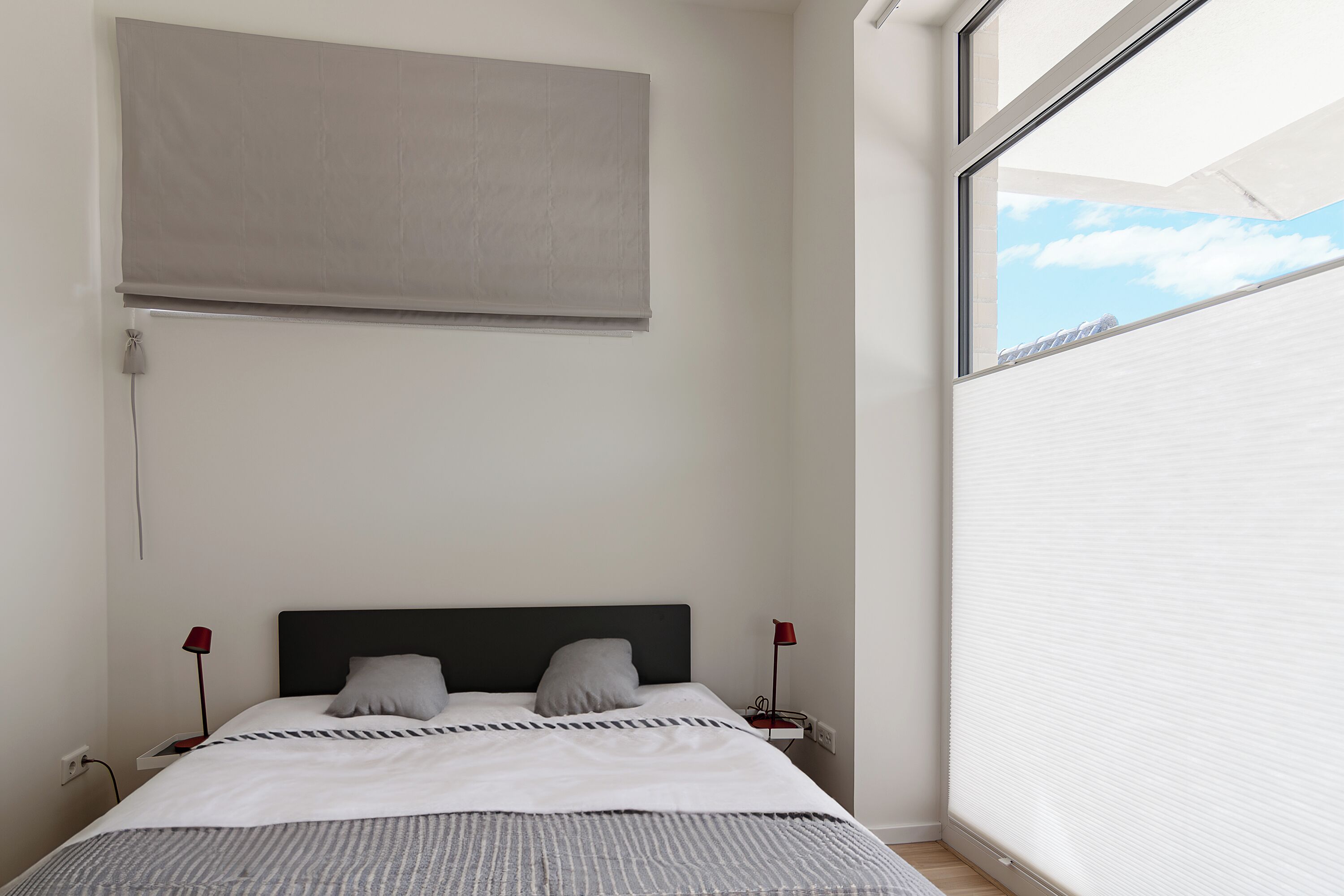 Ein Schlafzimmer mit einem Bett, zwei Nachttischlampen, einem Fenster mit Jalousien und einer Wandleuchte.