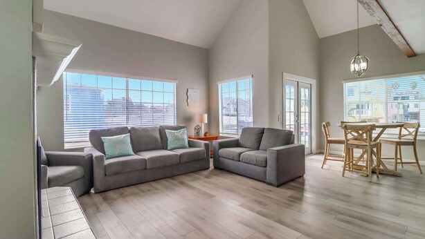 A spacious living room with a gray sectional sofa, a wooden dining table with chairs, and large windows with blinds. - Steps to bay & ocean, Belmont Park, A/C family fun (San Diego)