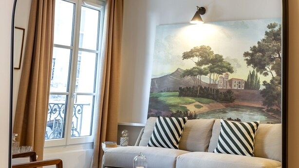 Un soggiorno con divano, poggiapiedi e tavolino, un quadro appeso alla parete e una finestra con tende.