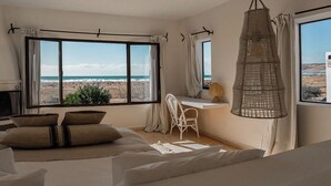A bedroom with a large window overlooking a beach, a bed with beige bedding, a white chair, and a hanging light fixture. - La Mouette et les Dromadaires (Essaouira)