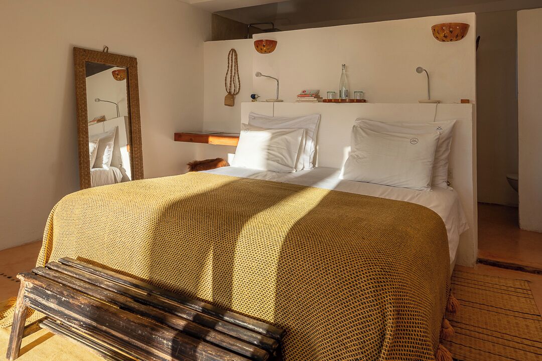 Un dormitorio con una cama grande, un banco de madera, un espejo, una mesita de noche y una estantería de pared con adornos.