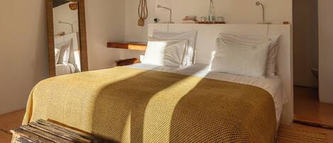 Un dormitorio con una cama grande, un banco de madera, un espejo, una mesita de noche y una estantería de pared con adornos.