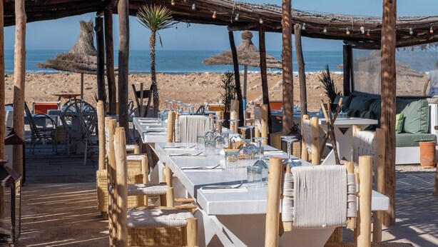 An outdoor dining area with wooden tables and chairs, a thatched roof, and a view of the beach and ocean. - La Mouette et les Dromadaires (Essaouira)