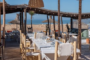 Un área de comedor al aire libre con mesas y sillas de madera, techo de paja y vistas a la playa y el océano.