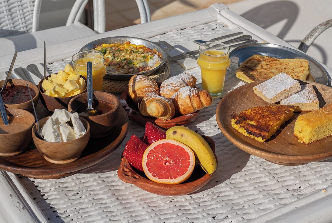 Un desayuno abundante sobre una mesa blanca al aire libre, con diferentes platos, frutas y un vaso de jugo de naranja.