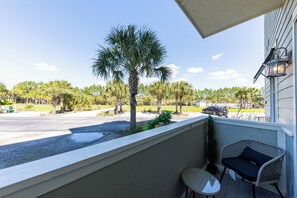 A balcony with a wicker chair and table, overlooking a street with palm trees and a parked car. - Stylish 30A Signature King Suite in Santa Rosa with Parking, Pool & Balcony (Santa Rosa Beach)