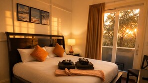 A hotel room with a bed, a nightstand with a lamp, a window with curtains, and a view of trees.