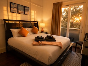 A hotel room with a bed, a nightstand with a lamp, a window with curtains, and a view of trees.