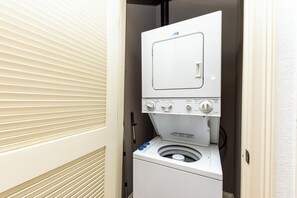 A compact, white laundry room with a built-in washer and dryer, a control panel, and a vent hose. - Stylish 30A Signature King Suite in Santa Rosa with Parking, Pool & Balcony (Santa Rosa Beach)