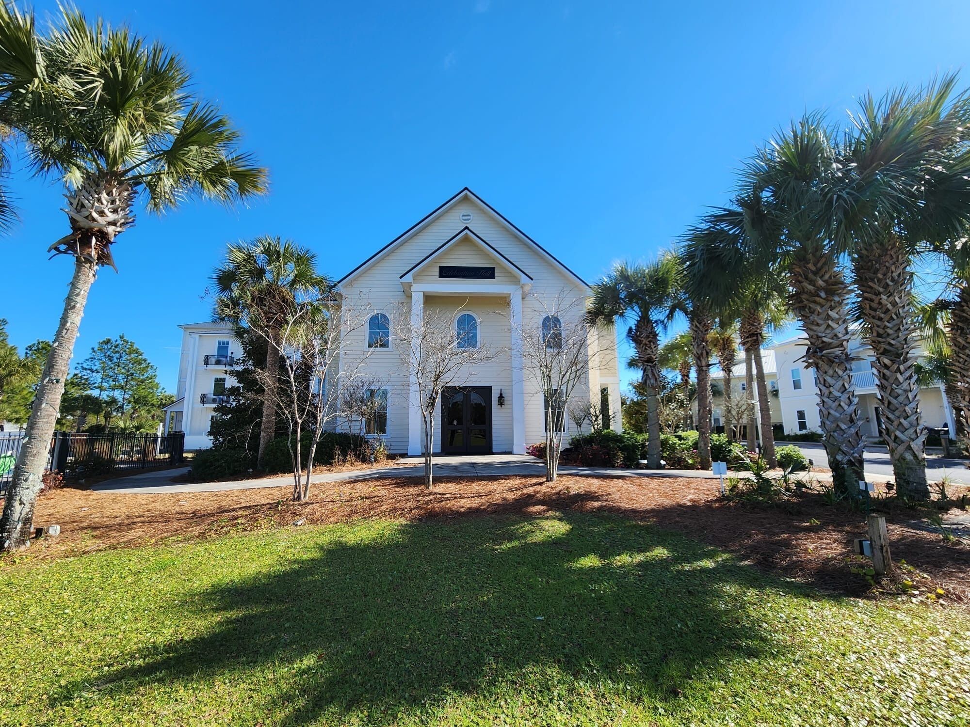 A two-story white building with a central entrance, surrounded by palm trees and a well-maintained lawn.