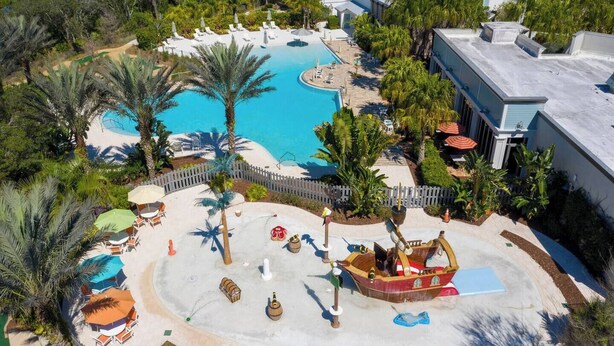 Vista aérea de un complejo turístico con piscina central, rodeado de palmeras y sillas de descanso.