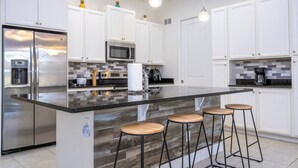 Una cocina moderna con armarios blancos, electrodomésticos de acero inoxidable, una isla grande con taburetes y un salpicadero con azulejos grises.