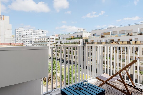 Ein Balkon mit Blick auf moderne Gebäude, einem Holztisch und einer Sonnenbrille.