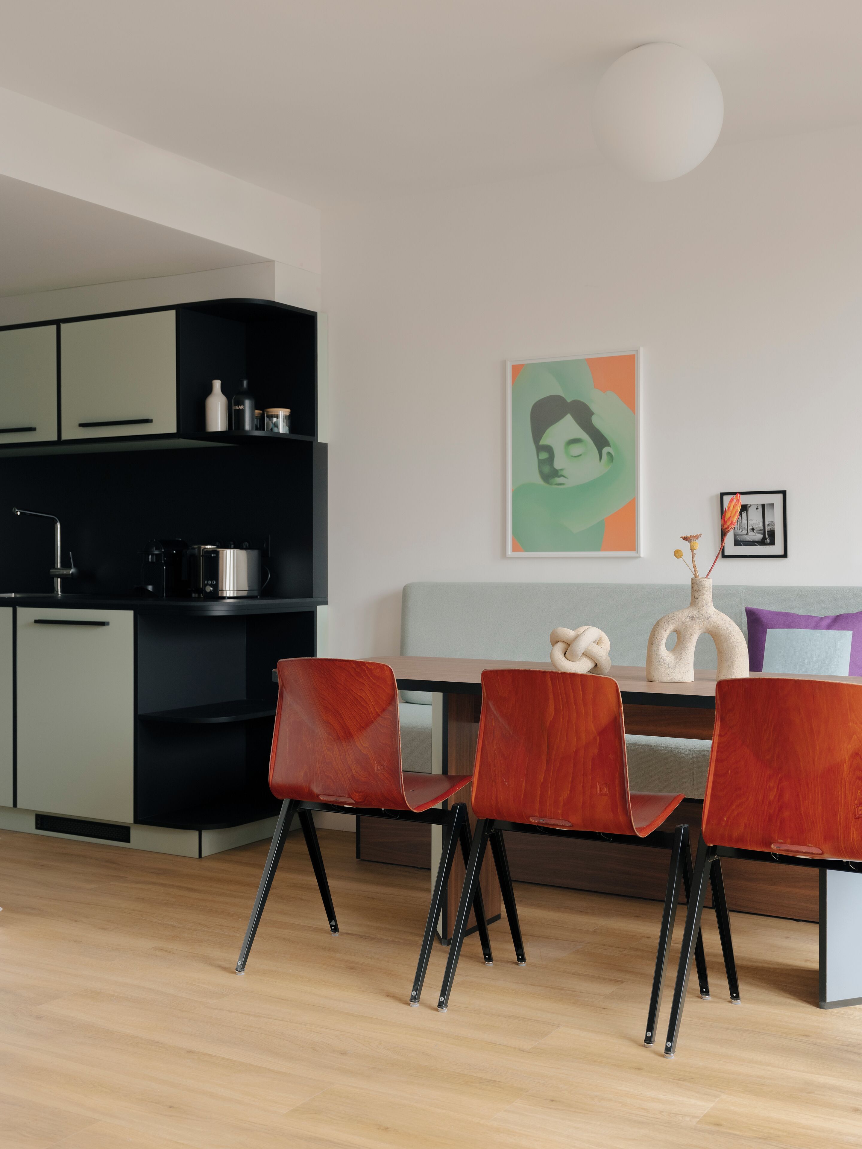 Een moderne keuken met zwarte kastjes, een houten eettafel en rode stoelen. Op de tafel staat een witte vaas met gedroogde bloemen.