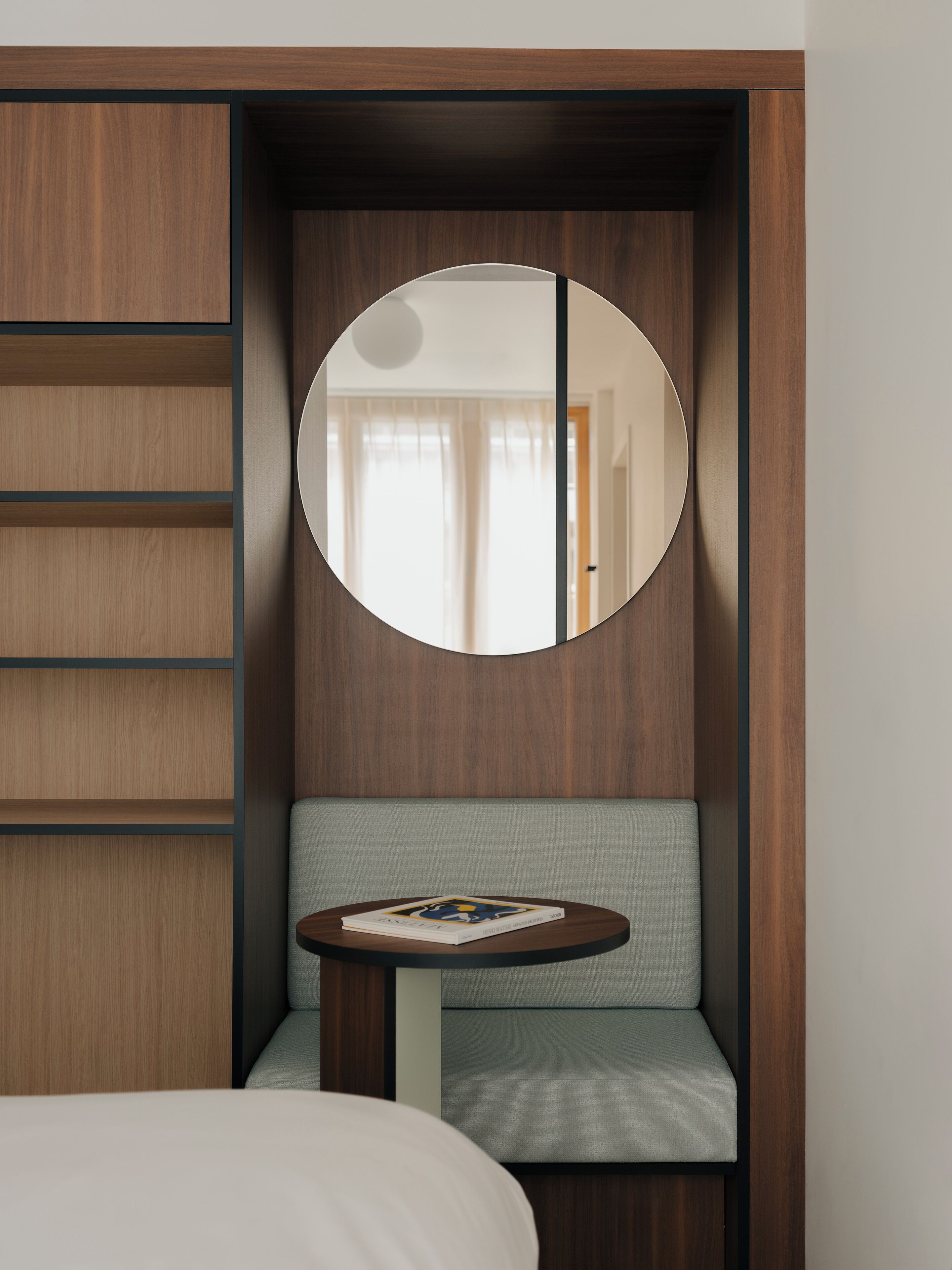 A hotel room with a wooden wardrobe, a round mirror, a grey sofa, and a small table with a magazine.