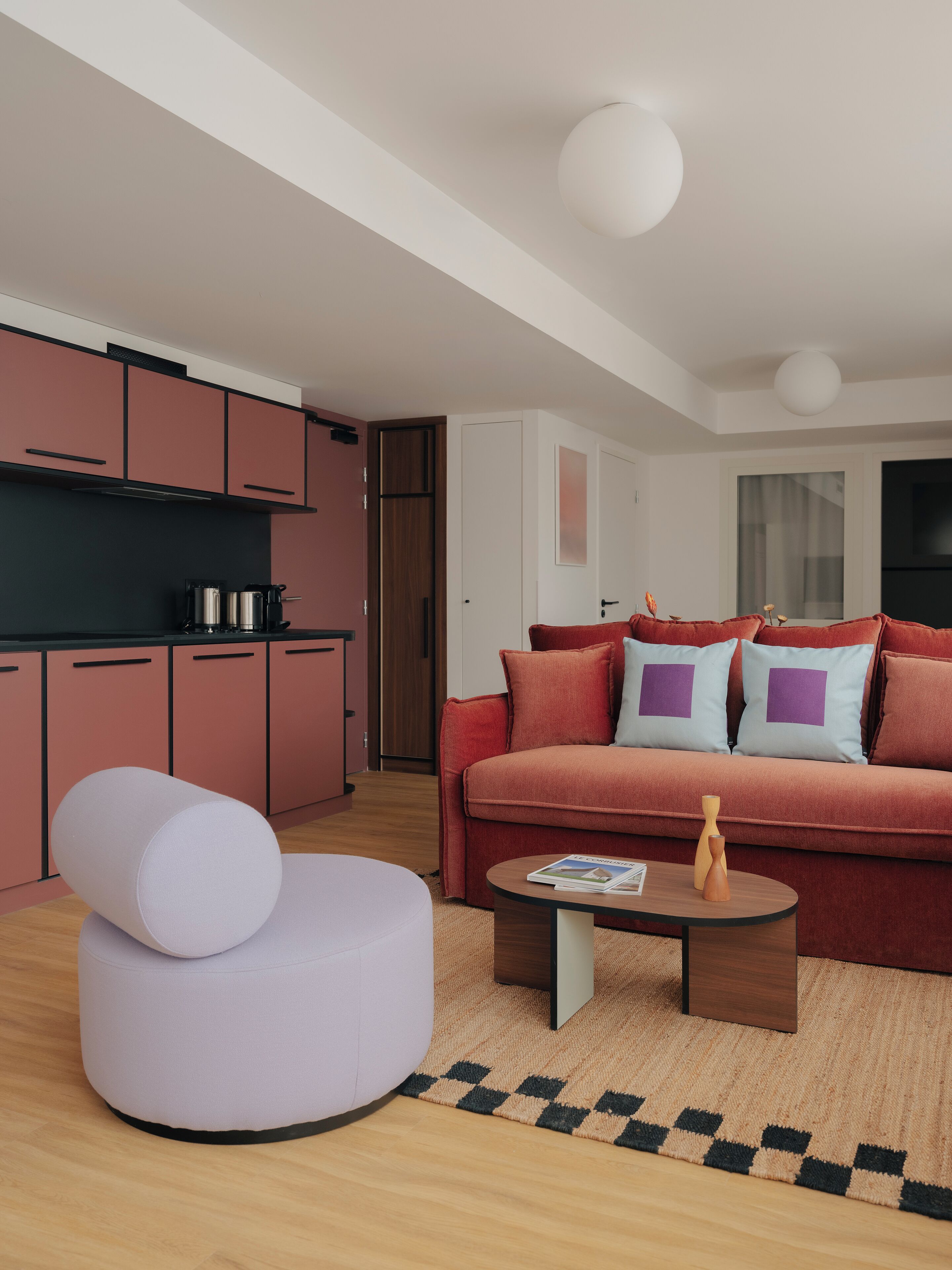 Ein modernes Wohnzimmer mit einem roten Sofa, einem weißen Hocker und einem Couchtisch. Es gibt eine Küchenzeile mit rosafarbenen Schränken und einer schwarzen Arbeitsplatte.
