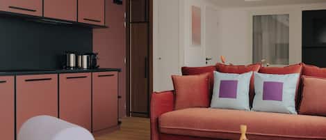 Una sala moderna con un sofá rojo, un reposapiés blanco y una mesa de centro. Hay una zona de cocina con armarios rosados y una encimera negra.